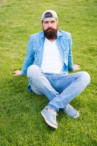 Zdecyduj się na prostotę nad showiness. Moda człowiek siedzi na zielonej trawie. Brodaty człowiek w modzie denim stylu na zewnątrz. Modny Hipster z długą brodą noszenie czapki mody do tyłu. Moda męska lato — Zdjęcie stockowe