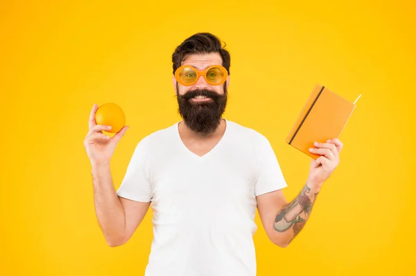 A good weight loss book. Bearded man holding book and orange fruit on yellow background. Hipster reading diet book. Keeping the book in home library