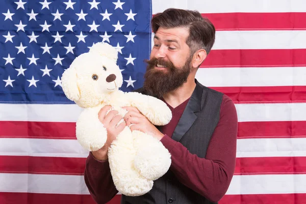 Július 4. Függetlenség napja. Amerikai srác a játék ünnepelni ünnep. Amerikai ünnep. Ember szakállas csípő tartsa mackó amerikai zászló háttérben. Gyermekgondozás. Még gyerekes. Csecsemő-és mulatságos — Stock Fotó