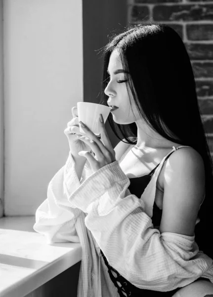Пийте добре почувайтеся. Гарна жінка п'є здоровий ранковий напій у вікні. Чарівна дівчина тримає чашку зі смачним кавовим напоєм. Сексуальна жінка насолоджується її рецептом напою — стокове фото