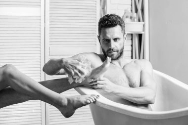 adam köpük sünger ile kadın bacaklarını yıkayın. maço banyo zevk. Banyodaki seksi adam. Seks ve rahatlama konsepti. Küvette maço çıplak. Köpükleri suyla dikkatlice yıkayın. kaslı vücutlu adam