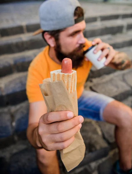 Guy eating hot dog. Man bearded bite tasty sausage and drink paper cup. Street food so good. Urban lifestyle nutrition. Carefree hipster eat junk food while sit on stairs. Hungry man snack. Junk food