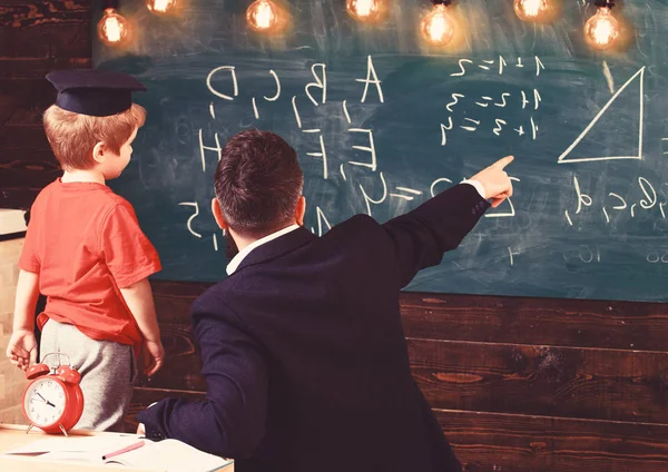 Unga manliga läraren vägleder sitt barn student till lärande medan peka och titta på tavlan med klotter på, sitter i klassrummet, bakifrån — Stockfoto