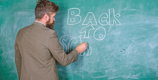 Man bearded teacher missed his work during vacation. Teacher near chalkboard holds chalk write inscription back to school. Back to school concept. Teacher passionate job ability reach out students