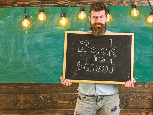 Teacher on suspicious face holds blackboard with title back to school. Man with beard welcomes students, chalkboard on background. Teacher peeking out of chalkboard. Start of school year concept