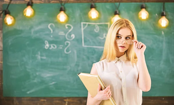 La mujer con libro comienza la lección, mira al público mientras se quita las gafas. El profesor se ve confiado en las gafas, de pie en el aula, pizarra en el fondo. Atractivo concepto de profesor — Foto de Stock