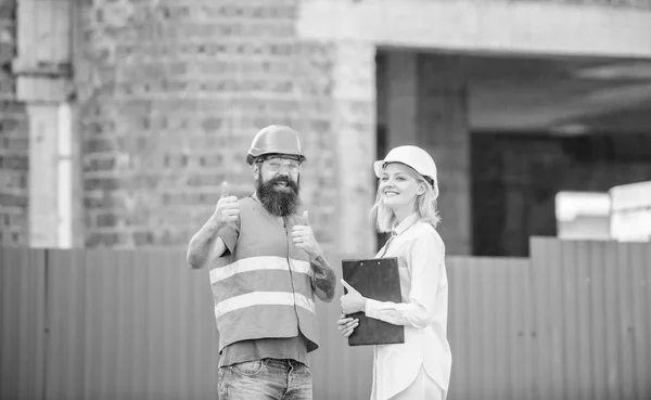 Discuss progress project. Woman inspector and bearded brutal builder discuss construction progress. Construction project inspecting. Safety inspector concept. Construction site safety inspection