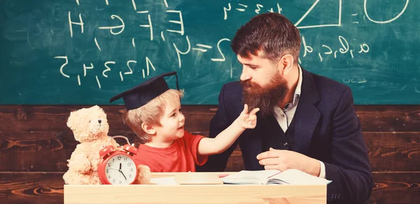 Stoute jongetje concept. Vader met baard, leraar leert zoon, jongetje. Kid vrolijke afleiden tijdens het studeren, aandacht tekort. Leraar en leerling in mortarboard, schoolbord op achtergrond — Stockfoto