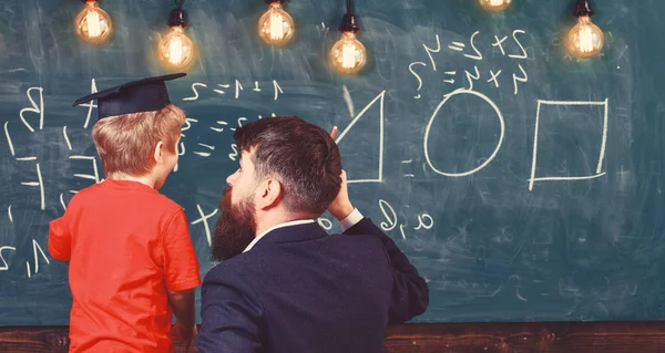 Ung lärare förklarar aritmetik till den lille pojken i Academic Cap. Man med skägg och blont barn vände tillbaka i klassrummet. Guy pekar på tavlan med former och siffror — Stockfoto