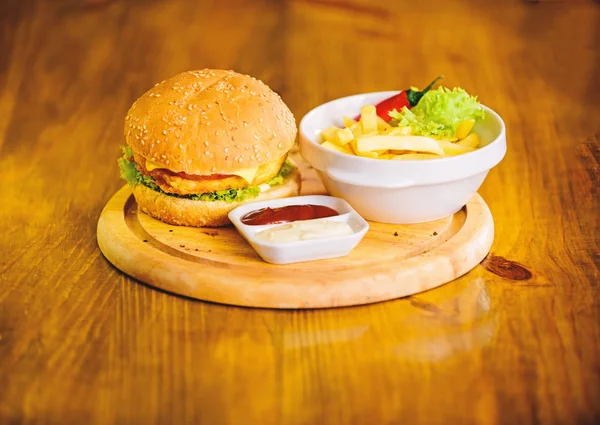 Hambúrguer com carne de queijo e salada. Comida de batota. Hambúrguer delicioso com sementes de sésamo. Conceito de fast food. Menu de hambúrgueres. Lanche de alta caloria. Hambúrguer e batatas fritas e molho de tomate em tábua de madeira — Fotografia de Stock