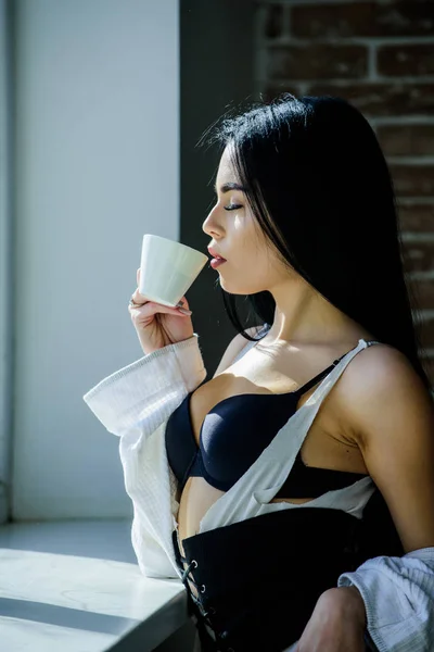 Njuter av morgonsolen. Sensuell tjej sexiga bröst dricka kaffe nära fönsterbrädan. Attraktiva kvinnliga underkläder som kopplar av hemma. Passionerad mystisk älskare. Perfekt kaffe tid. Men först kaffe — Stockfoto