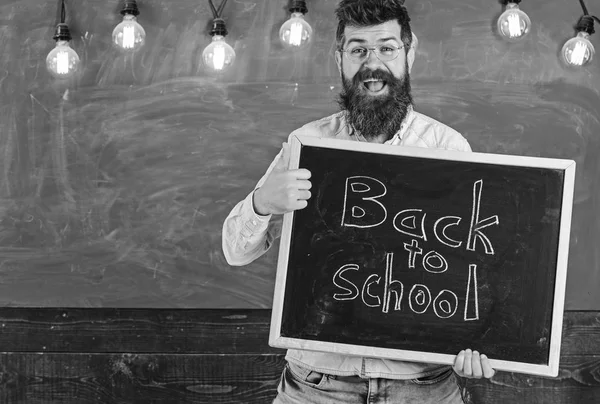 Вчитель в окулярах тримає дошку з написом назад до школи. Чоловік з бородою на щасливому обличчі вітає студентів з великим пальцем вгору жестом, крейдою на фоні. Повернутися до концепції школи — стокове фото