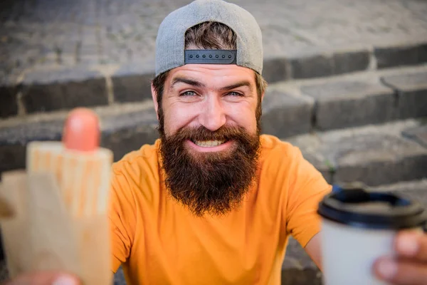 Man bearded eat tasty sausage and drink paper cup. Urban lifestyle nutrition. Junk food. Carefree hipster eat junk food while sit stairs. Snack for good mood. Guy eating hot dog. Street food concept