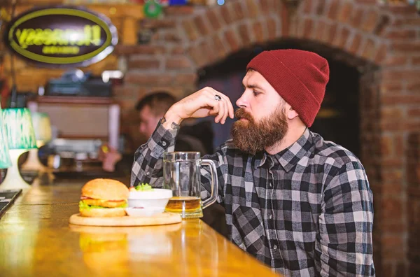 High calorie snack. Brutal hipster bearded man sit at bar counter. Hipster relaxing at pub. Pub is relaxing place to have drink and relax. Man with beard drink beer eat burger menu. Enjoy meal in pub