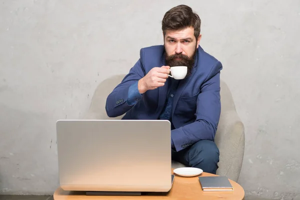 In mobile style. Bearded man enjoying his hot drink and mobile internet. Businessman with mobile device drinking tea or coffee at workplace. Hipster in formalwear working at laptop in mobile office