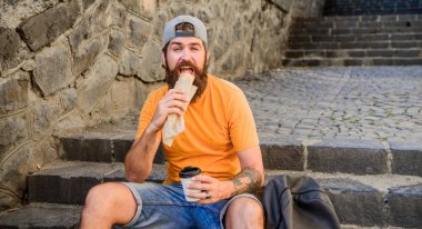 Adam sakallı hızlı atıştırmalık ve içecek kağıt fincan zevk. Sokak Yemekleri çok güzel. Kentsel yaşam tarzı beslenme. Merdivenlerde oturup Carefree hipster önemsiz yiyecek yemek. Aç adam atıştırmalığı. Önemsiz yiyecekler. Adam yemek sıcak köpek