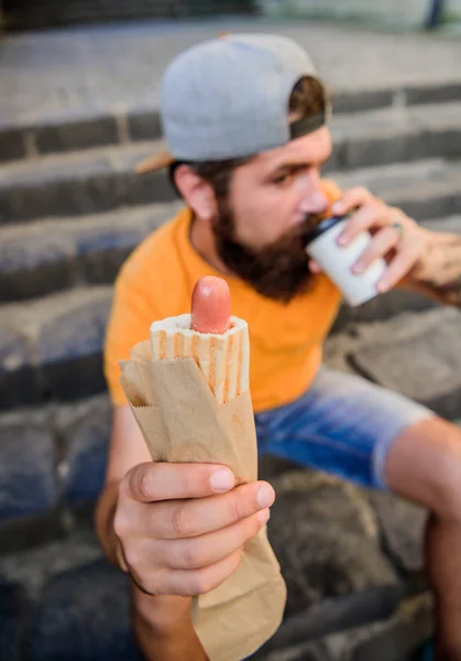 Man bearded bite tasty sausage and drink paper cup. Street food so good. Urban lifestyle nutrition. Carefree hipster eat junk food while sit on stairs. Hungry man snack. Junk food. Guy eating hot dog
