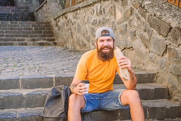 Comida deliciosa para o seu estilo de vida. Viajante caucasiano gosta de comida de rua. Um homem barbudo a comer comida insalubre. Hipster descansando e tendo fast food cachorro quente durante a viagem de verão — Fotografia de Stock