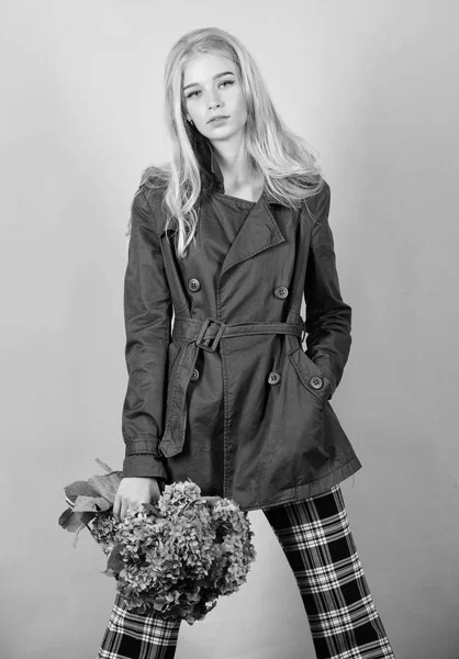 Trench coat fashion trend. Must have concept. Fashionable coat. Woman blonde hair posing coat with flowers bouquet. Clothes and accessory. Girl fashion model wear coat for spring and autumn season