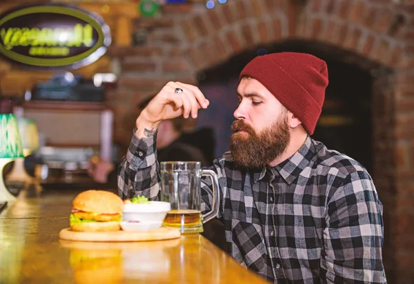 High calorie snack. Hipster relaxing at pub. Pub is relaxing place to have drink and relax. Man with beard drink beer eat burger menu. Enjoy meal in pub. Brutal hipster bearded man sit at bar counter