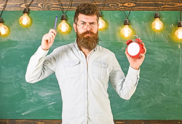 Teacher in eyeglasses holds alarm clock and pen. Discipline concept. Bearded hipster holds clock, chalkboard on background. Man with beard and mustache on strict face stands in classroom — Stock Photo, Image