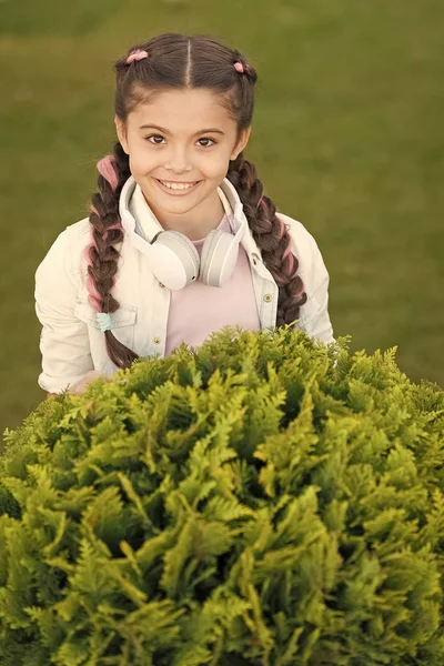 Segredos para criar uma criança feliz. Menina bonito miúdo verde grama fundo. Criança feliz emocional saudável relaxando ao ar livre. O que faz a criança feliz. Tranças menina penteado e fones de ouvido modernos desfrutar relaxar — Fotografia de Stock