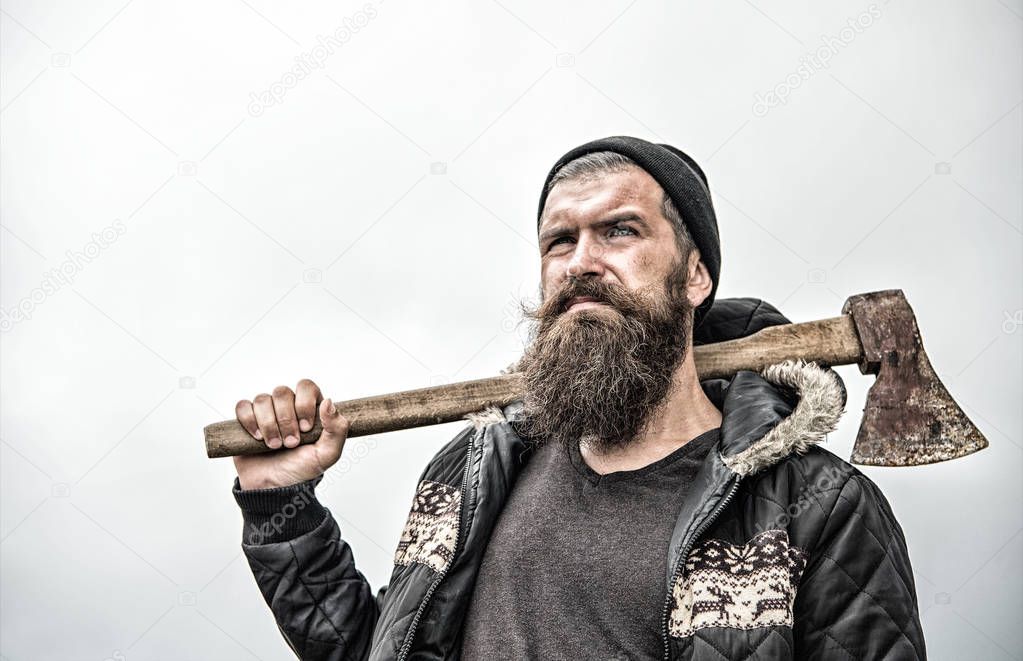 Hipster with beard on serious face carries axe on shoulder sky on background, copy space. Lumberjack brutal and bearded holds axe. Brutal lumberjack concept. Man in hat and warm jacket looks brutally
