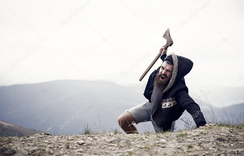 Man with beard on shouting face conquers top of mountain with axe, sky background. Survive in wild nature concept. Hipster with axe surviving in mountains. Guy brutal and bearded in wild nature