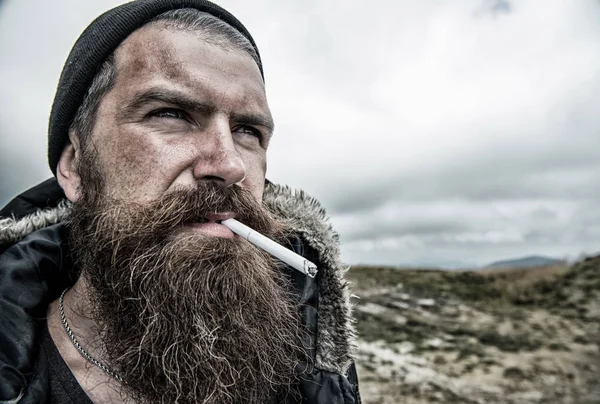 Man with long beard and mustache smoking cigaret. Brutality concept. Hipster on strict face with beard looks brutally while hiking and smoking. Man with brutal bearded appearance, untidy and unshaven