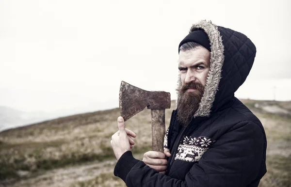 Hipster med skägg på strikt ansikte håller skarp yxa, skyline på bakgrund, kopia utrymme. Lumberjack brutala och skäggiga innehar axe. Brutala lumberjack koncept. Man i hatt och varm jacka ser brutalt — Stockfoto