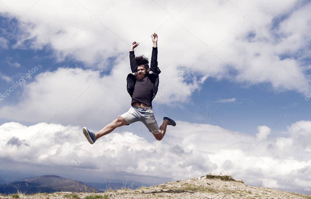 Hipster or brutal macho conquers mountain. Man with brutal appearance jumps. Freedom concept. Man with beard enjoy freedom, jumpimg on top of mountain. Hipster feels free while hiking, sky background