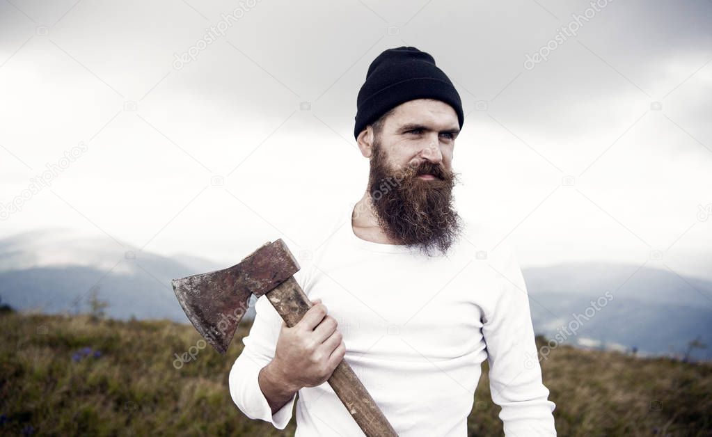 Hipster with beard on strict face holds axe, skyline on background. Lumberjack brutal and bearded holds axe while stand on top of mountain. Hiking concept. Hipster looks brutal and stylish with axe