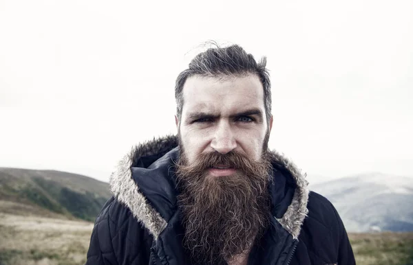 Man with long beard and mustache wears jacket. Hipster on strict face with beard looks brutally while hiking. Hermit concept. Man with brutal bearded appearance, brutal unshaven man looks untidy