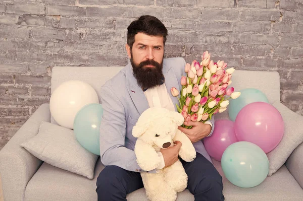 Romantic man with flowers and teddy bear sit on couch waiting girlfriend. Romantic gift. Macho getting ready romantic date. Man wear blue tuxedo bow tie hold flowers bouquet. Best boyfriend ever