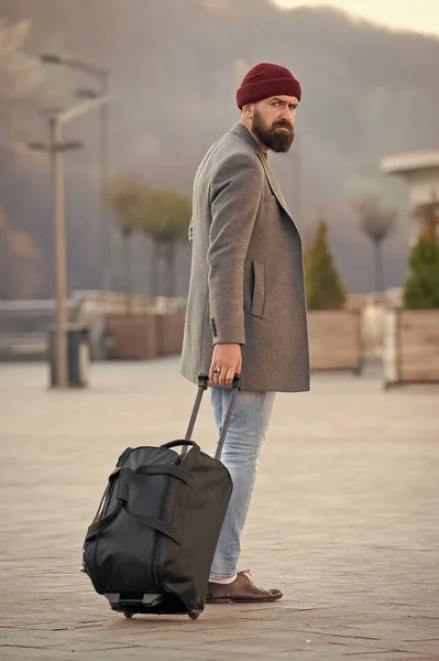 Carry travel bag. Man bearded hipster travel with luggage bag on wheels. Adjust living in new city. Traveler with suitcase arrive airport railway station urban background. Hipster ready enjoy travel