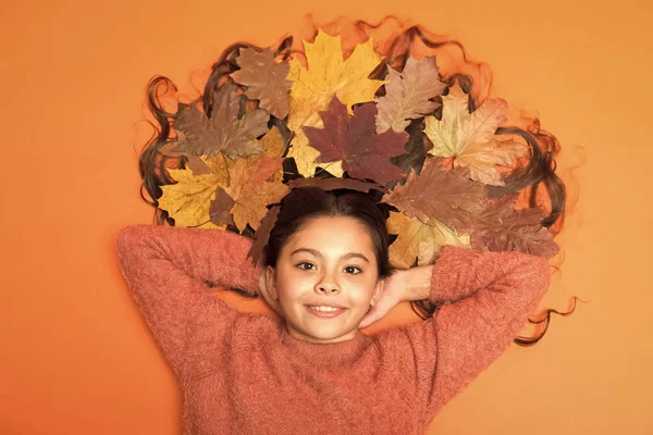 Der Sturz ist ihr in den Sinn gekommen. Mädchen niedliches Kind lag auf orangefarbenem Hintergrund mit abgefallenen Blättern. Kinder genießen die Herbstzeit. Trockene Ahornblätter in ihrer Frisur. Herbstzeitkonzept. Haarpflege im Herbst Tipps und Ideen — Stockfoto