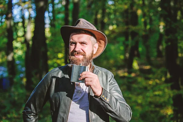 Hombre hipster relajarse en el bosque de otoño. Primavera tiempo soleado. camping y senderismo. macho maduro con aspecto brutal beber alcohol de matraz metálico. Hombre barbudo con sombrero de vaquero en el parque al aire libre. Vaquero sonriente —  Fotos de Stock