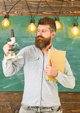 Gözlük öğretmen kitap ve mikroskop tutar. Sakal ve bıyık sınıfta sıkı yüzünde olan adam. Bilim adamı kitap ve mikroskop, kara tahta arka plan üzerinde tutar. Bilimsel araştırma kavramı