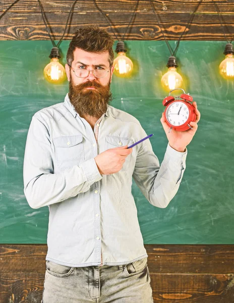 Professor em óculos detém despertador e caneta. O hipster barbudo segura relógio, quadro-negro no fundo. Homem com barba e bigode no rosto estrito fica na sala de aula. Conceito de disciplina — Fotografia de Stock