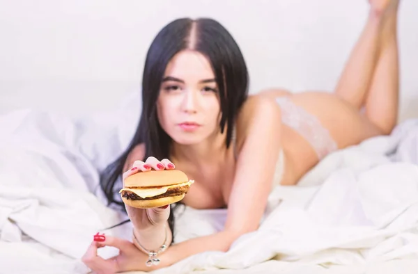 Dziewczyna w seksownej bieliźnie jeść Burger. Seksualny apetyt. Usługi dostarczania żywności. Uwodzicielski sexy kobieta zrelaksować się na łóżku. Koncepcja diety. Fast food. Uwodzicielski kobieta głodny Leniwe jedzenie dnia. Jedzenie śmieci w łóżku — Zdjęcie stockowe