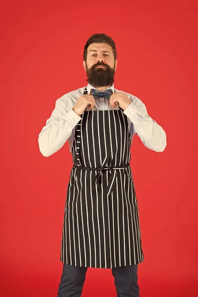 His style works really well to compliment beard. Bearded man fixing bow tie in bib apron. Elegant hipster with bearded face. Bearded bartender or cook in work uniform. Long bearded waiter or servant