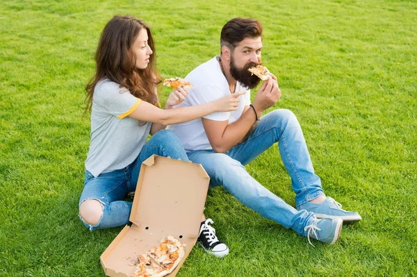 Couple eating pizza relaxing on green lawn. Fast food delivery. Bearded man and woman enjoy cheesy pizza. Couple in love dating outdoors with pizza. Hungry students sharing food. Pure enjoyment