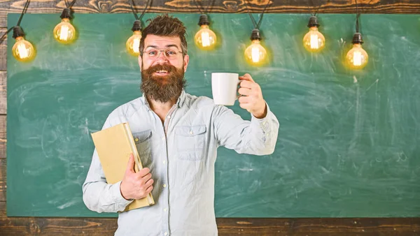 Kávészünet koncepció. Férfi szakáll boldog arc az osztályteremben. Tanár a szemüvegek tartja a könyvet, és a bögre kávét vagy teát. Tudós tartja a könyvet, és a bögre kávét, chalkboard háttér, másolás spac — Stock Fotó