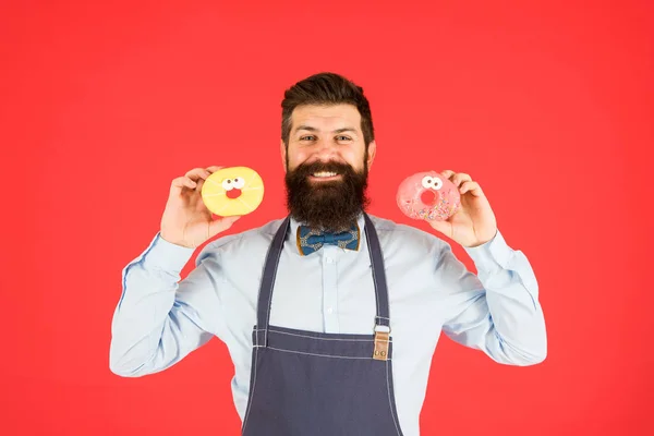 Boulangerie. Des pâtisseries. Bonbons et gâteaux. De la malbouffe. Hipster barbu boulanger tenir des beignets. Boulangerie de beignets. Un beignet glacé. Barbu gai homme bien entretenu dans un tablier de vente de beignets. Propriétaire de boulangerie — Photo