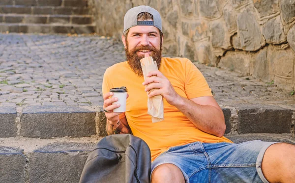 Comida de plástico. Um tipo a comer cachorros quentes. Homem barbudo desfrutar de lanche rápido e beber copo de papel. Comida de rua tão boa. Nutrição de estilo de vida urbano. Despreocupado hipster comer junk food while sit on stairs. Lanche de homem faminto — Fotografia de Stock