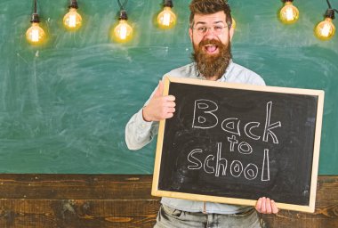 Gözlüklü öğretmen okula geri yazıt ile tahta tutar. Mutlu yüzünde sakallı adam başparmak yukarı jest, arka planda tebeşir ile öğrencileri ağırlıyor. Okula geri dönüş concep