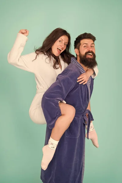 Couple in bathrobes having fun turquoise background. Lets stay at home and have fun. They always have fun together. Enjoying every second together. Handsome young man piggybacking beautiful woman