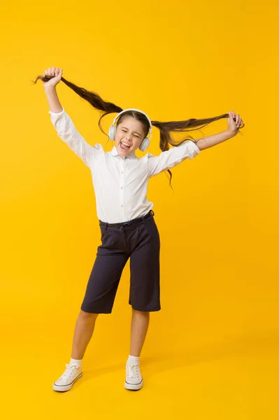 Urosną włosy z muzyką. Małe dziecko noszące długie ogonowe fryzury i słuchawki do szkoły. Mała dziewczynka trzyma fryzurę na żółtym tle. Cute kid korzystających z muzyki i jej fryzury. Łatwa fryzura — Zdjęcie stockowe