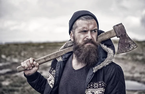 Bearded man hold rusty axe on natural background. Hipster in hat and jacket outdoor. Man with beard and moustache on serious face. Discovery and adventure. Camping and wanderlust, vintage filter