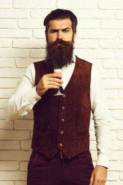 Uzun sakallı ve bıyıklı yakışıklı adamın ciddi yüzünde şık saçlar var. Beyaz tuğla duvar arka planında klasik süet deri yelek giymiş alkolik bardakları tutuyor.
